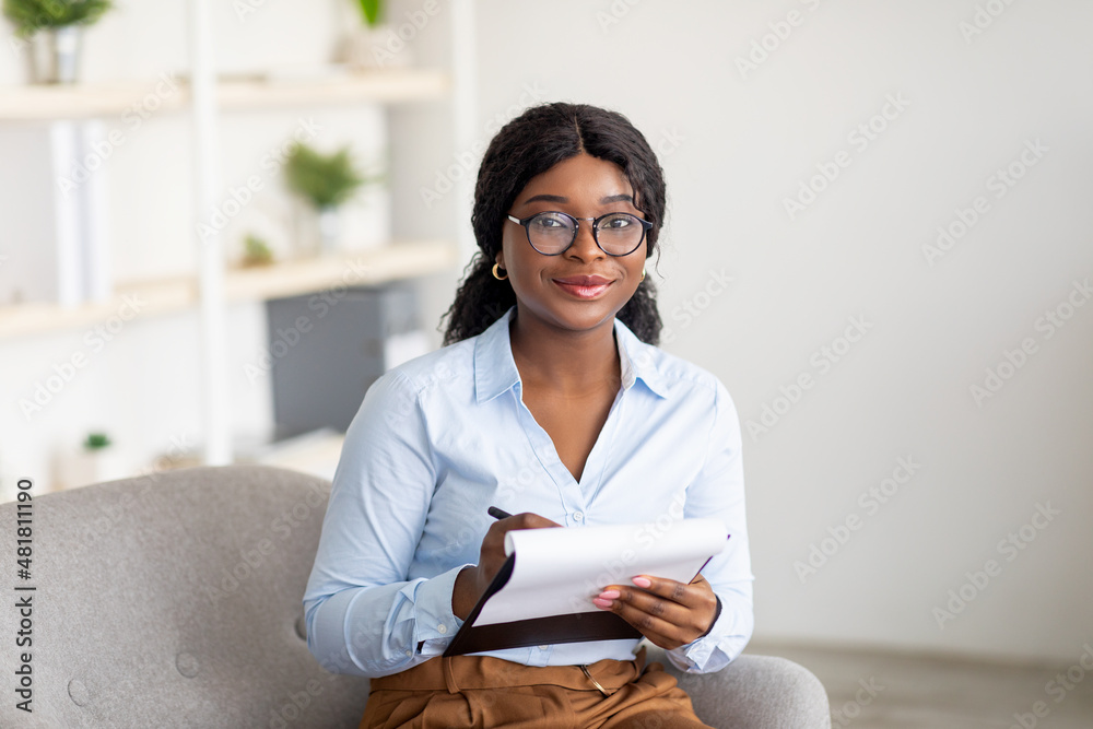 Friendly black female psychologist writing in clipboard, having session with client, sitting on couch at modern office - obrazy, fototapety, plakaty 