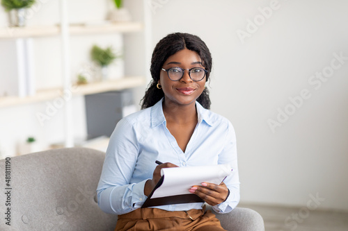 Friendly black female psychologist writing in clipboard, having session with client, sitting on couch at modern office