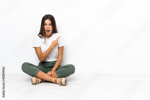 Teenager girl sitting on the floor surprised and pointing side