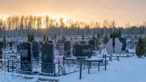Fotografie, Obraz Winter sunset over a snow covered cemetery with dark headstones and trees
