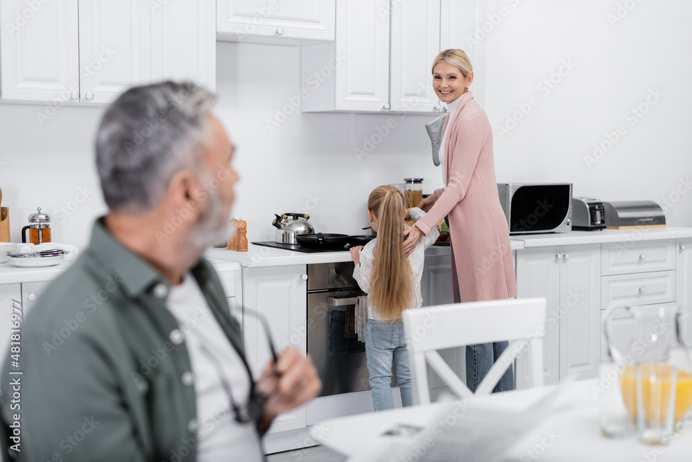 Obraz premium smiling woman looking at blurred husband while preparing breakfast with granddaughter.