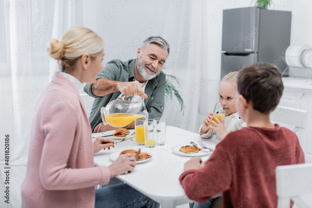 senior man pouring orange juice during breakfast with wife and grandchildren.