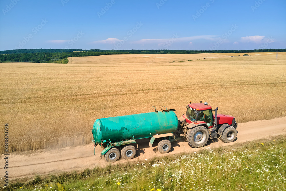 Obraz premium Tractor with water tank trailer driving on dirt road between agricultural fields during harvest season