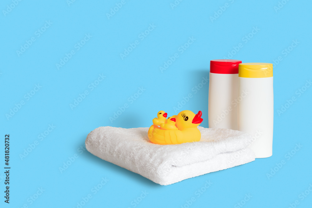 Yellow rubber duck on soft white towel and two plastic white bottles with yellow and red caps on light blue background. Child bath accessories, baby care. Copy space.