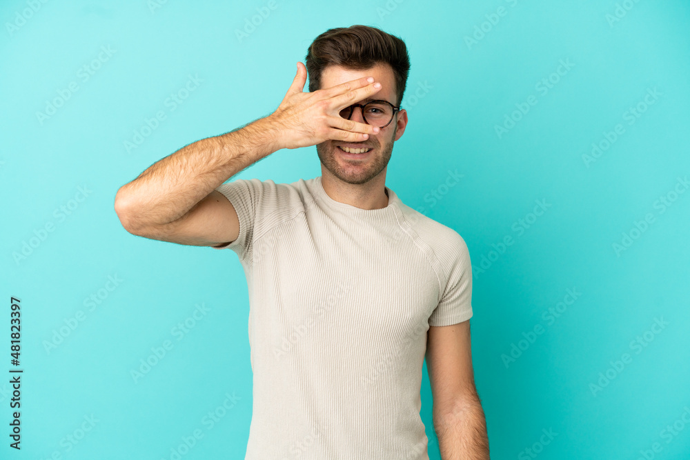 Fototapeta premium Young caucasian handsome man isolated on blue background covering eyes by hands and smiling