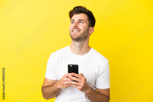 Young caucasian handsome man isolated on yellow background using mobile phone and looking up © luismolinero