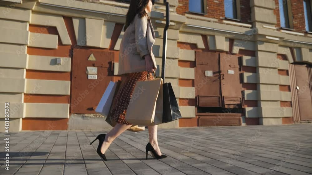 Fashionable woman carries paper packages going along urban street. Young girl holds shopping bags walking at sidewalk after purchases. Concept of seasonal sales and discounts. Dolly shot Slow motion