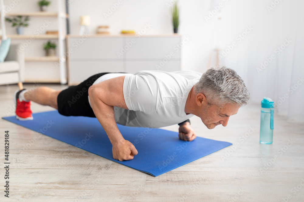 Active mature man doing push-up exercises on fists, training on mat in living room at home, copy space
