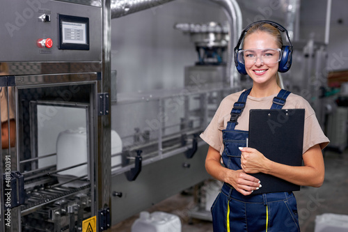 Female worker in protective workwear with folder-tablet working in medical supplies research and pesticides production factory, controlling production of bottles canisters before shipment, smile