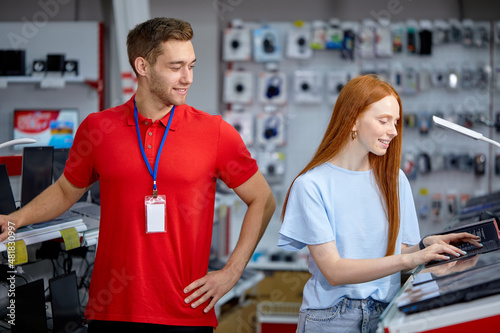 redhead woman is looking for the best laptop in computer department of electronics store. Caucasian buyer chooses laptop in modern technology store, pleasant male consultant help customer