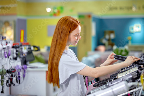 Pleasant redhead woman with long hair,choosing hair straightener in store. Female is standing by shelves with household appliances. The concept of buying household appliances for beauty and self-care