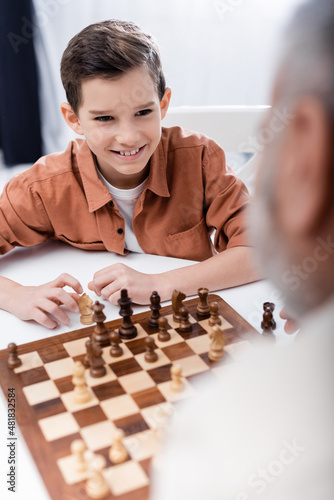 cheerful boy playing chess with blurred grandpa at home.