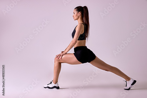 Portrait of young brunette lady stretching legs before workout, isolated in studio on pink background. Fit athlete female in black top and shorts warming up legs muscles, doing lunges
