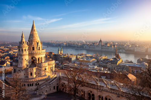 The main tower of the impressive Fisherman's Bastion (Halaszbastya) from above with Hungarian Parliament building and River Danube at background during a golden sunrise in Budapest photo