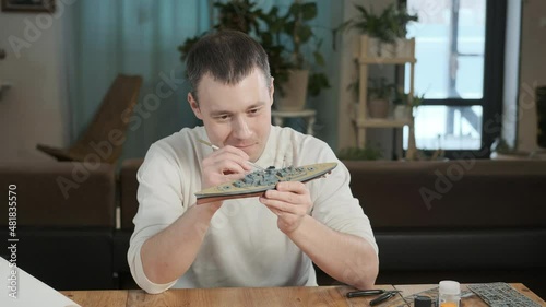 Happy adult male assembler plastic model of warship according to drawings and instructions.  man paints elements of layout with a brush. embodiment of childhood dream. Large-Scale Modeling