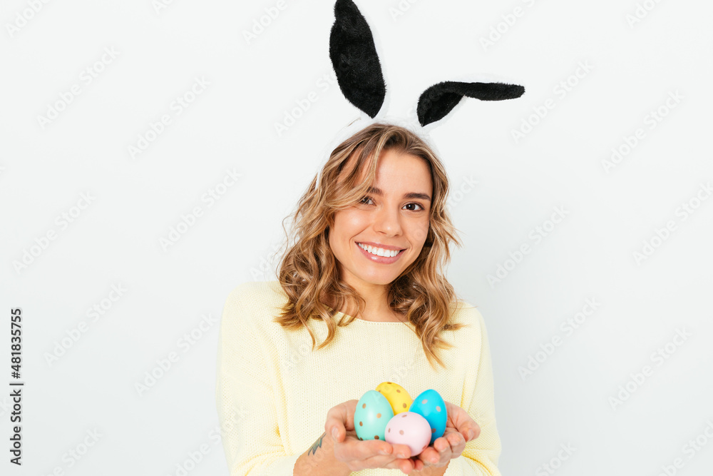 Lovely happy young woman celebrating Easter wears bunny ears holding several colored eggs Lovely happy young woman celebrating Easter wears bunny ears holding several colored eggs