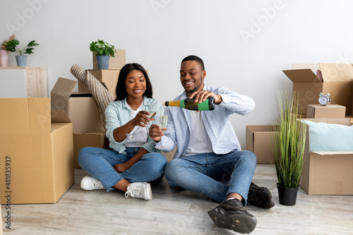 Loving black spouses celebrating moving day with champagne, sitting on floor in new apartment among cardboard boxes © Prostock-studio