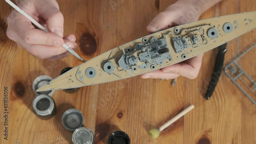 Passion for modeling and building toys. Close-up of man painting elements of layout with brush. Happy male assembler plastic model of warship according to drawings and instructions. Scale Modeling