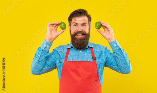 Fotografie Happy man in red apron holding fresh limes citrus fruits yellow background, gree