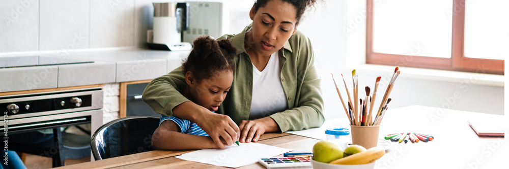 Black mid woman drawing with her focused daughter at home kitchen Black mid woman drawing with her focused daughter at home kitchen