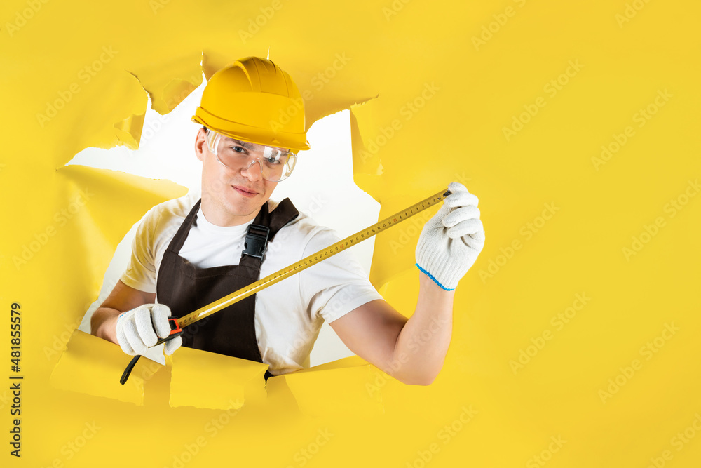 Confident mechanic, construction man. Happy carpenter. Handyman is measuring size of torn hole in yellow background. Senior builder engineer is holding tape-line for measure. Engineering and repair. Confident mechanic, construction man. Happy carpenter. Handyman is measuring size of torn hole in yellow background. Senior builder engineer is holding tape-line for measure. Engineering and repair.