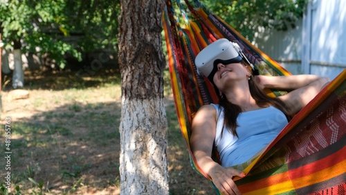 joyful woman wearing virtual reality glasses rests in a hammock near a tree, imagining a dream come true, an invented world. 