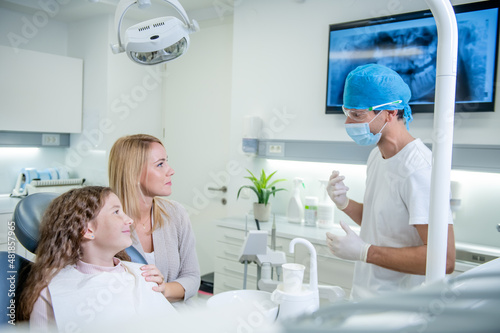 Middle shot of a male Caucasian dentist with protective gloves, face mask, and surgical cap advising and showing to a patient, a female child, and her mother an X-ray of the teeth on the screen. photo
