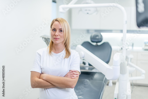 Middle shot of a Caucasian woman wearing a white T-shirt, standing in front of a dental chair at a dental clinic. © TheSupporter