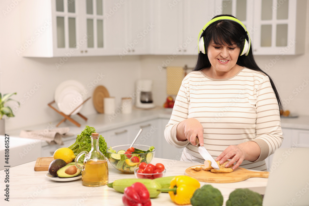 Happy overweight woman with headphones preparing healthy meal at table in kitchen