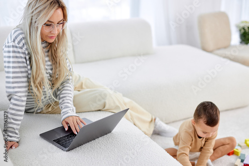 Bilde på lerret blonde caucasian mom working at home on sofa writing an email while child playing on floor