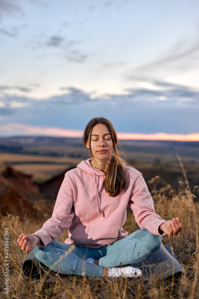 Young Female Keep Calm Sitting On Fitness Mat, Meditating With Eyes Closed Alone On Fresh Air. Attractive Young Lady In Sportive Leggins And Pink Hoodie relaxing, Having Rest. In Nature In Morning