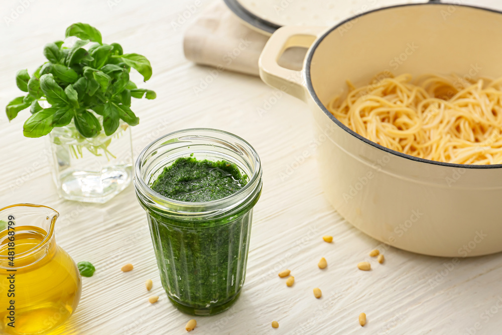 Jar of tasty pesto sauce, cooking pot of pasta and ingredients on light background Jar of tasty pesto sauce, cooking pot of pasta and ingredients on light background