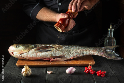Professional chef prepares silver carp herring in the restaurant kitchen. The cook sprinkles pepper on the fish. Work environment on the kitchen table.