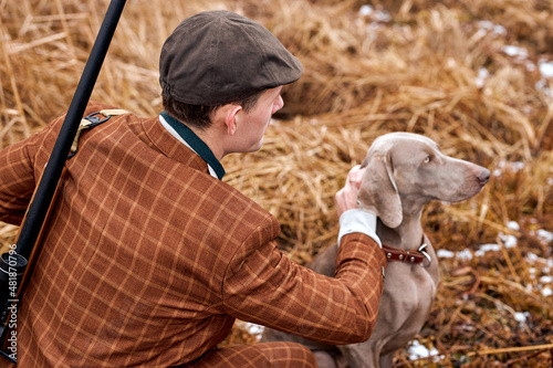 rear view on male with gray dog sitting in field waiting for target wild animal trophy, young guy in brown formal suit in nature, with shotgun. hunting, activ lifestyle, hobby concept