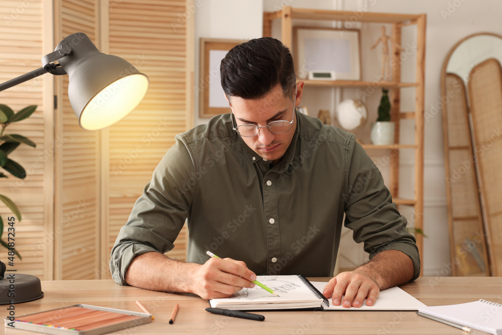 Man drawing in sketchbook with pencil at wooden table indoors