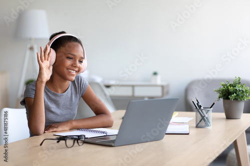 Smiling teen african american lady student in headphones studying at home with laptop  calling online and waving hand