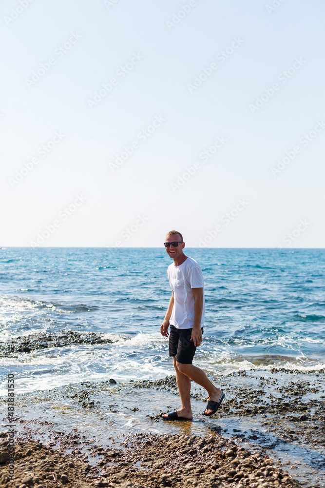 Young attractive man in sunglasses in a white t-shirt and shorts stands on the shore of the mediterranean sea