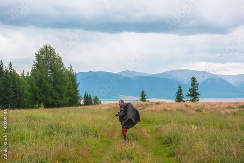 Satisfied tourist with camera in mountains in overcast. Happy man in raincoat walks through hills and forest in bad weather. Traveler goes towards adventure. Hiker and mountain range under cloudy sky.