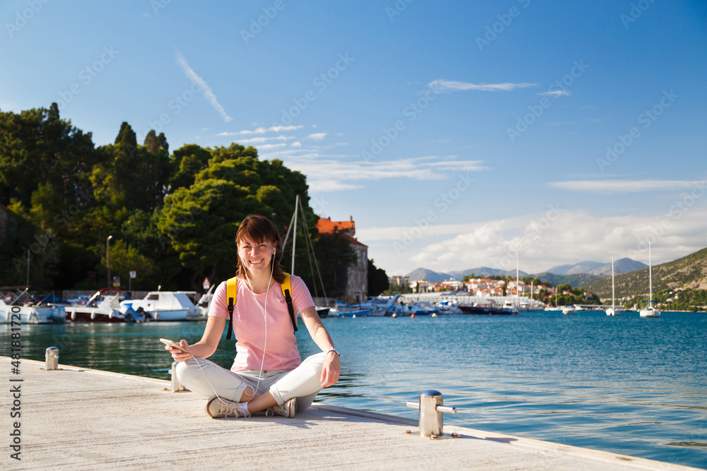 Smiling young female tourist sitting cross-legged on pier holding mobile phone with Croatian coastline in background. Bright sunny day in Dubrovnik, Croatia. Travel lifestyle concept