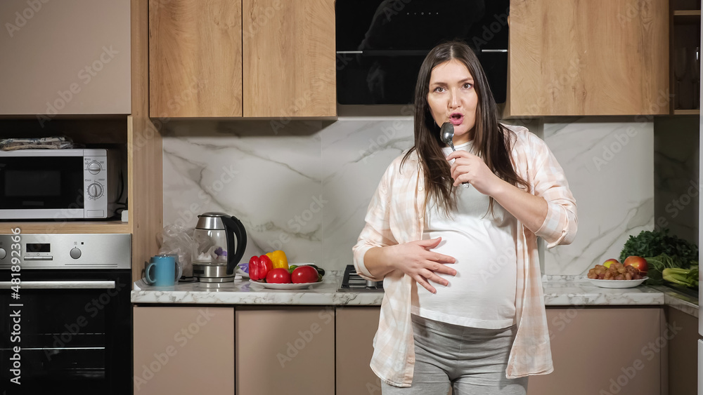 Pregnant woman with long brunette hair in comfortable domestic clothes dances and sings with spoon imitating microphone in kitchen near counters.