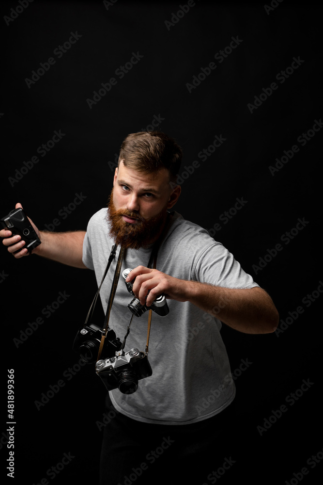 Handsome bearded photographer in a grey t-shirt with a bunch of different cameras in a hands and on a shoulder looking on a camera and ready for make a good shoot. Handsome bearded photographer in a grey t-shirt with a bunch of different cameras in a hands and on a shoulder looking on a camera and ready for make a good shoot.