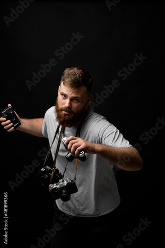 Handsome bearded photographer in a grey t-shirt with a bunch of different cameras in a hands and on a shoulder looking on a camera and ready for make a good shoot.