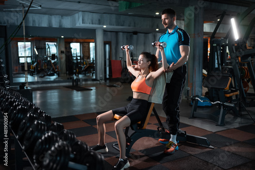young female exercising in gym with dumbbell with personal athletic trainer