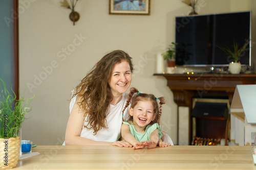 Portrait happy smile family. Mom and daughter at table look at cameras. two sisters laugh At home room and laugh. Place for text. childhood. Caucasian woman and girl in frame. Nanny child Babysitter.