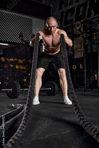 Sport. Battle ropes session. Attractive young fit caucasian sportsman working out in functional training gym doing exercise with battle ropes. In sports club. Portrait, single shirtless male