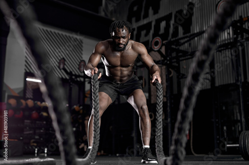 afro american fit sportsman doing battle ropes exercise at crossfit gym. African Man wear black shorts training with rope. sport motivation  cross fit  fitness concept. cardio training