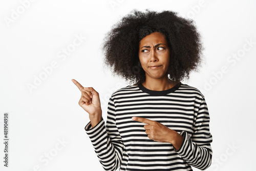 Image of young african american woman look with doubt, pointing and staring left with disappointed, skeptical frowning face, white background