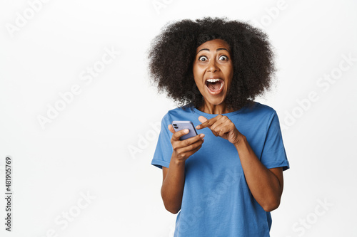 Image of african american modern woman in blue t-shirt, reacting amazed and surprised at mobile phone app, announcement on smartphone, smiling happy and excited