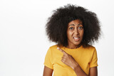Close up portrait of hesitant lady in yellow t-shirt, pointing finger left and asking question, being unsure, doubtful in smth, standing over white background