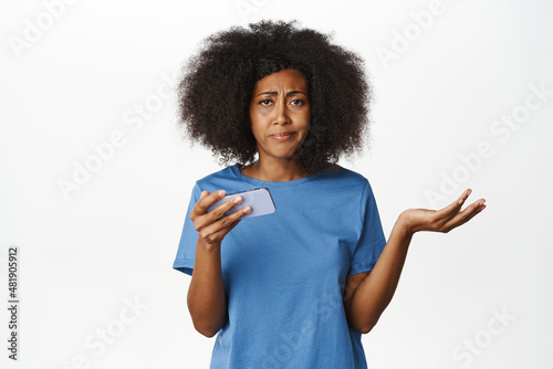 Confused african american woman shrugging, holding smartphone, staring puzzled at camera, standing in blue t-shirt over white background